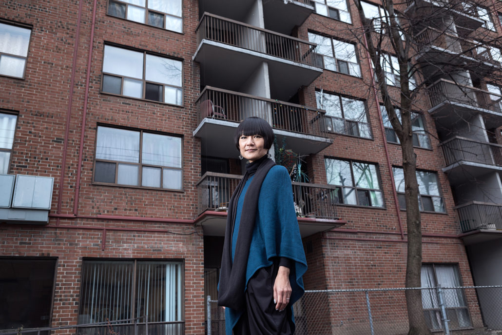 Toronto housing advocate Aiko Ito wearing blue sweater and black scarf in front of brick apartment building
