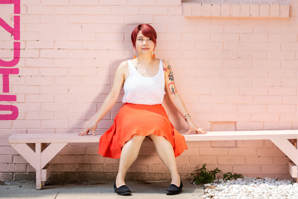 Van Loc in white top orange skirt seated before pink wall