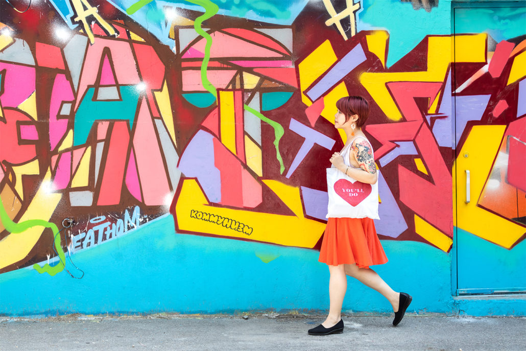 Van Loc in orange skirt walking before multi colour graffiti by Fathom