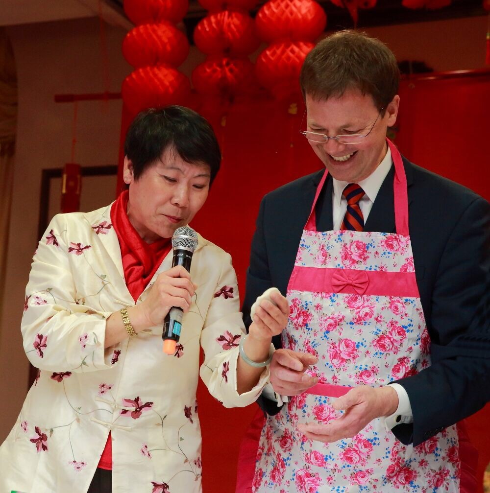 Nelly Gong holds a microphone and stands next to a man in an apron at a community event. 