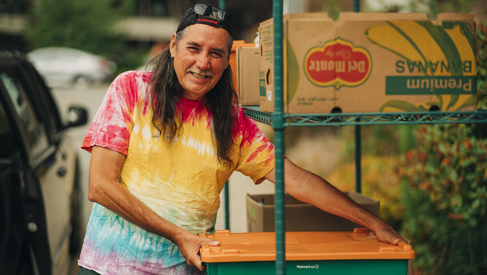 Chad wears a tie-dye t-shirt and baseball cap as he loads a box of groceries onto a shelf.  