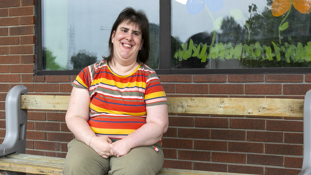 Cindy wears a striped t-shirt and sits on a couch in a rec room.