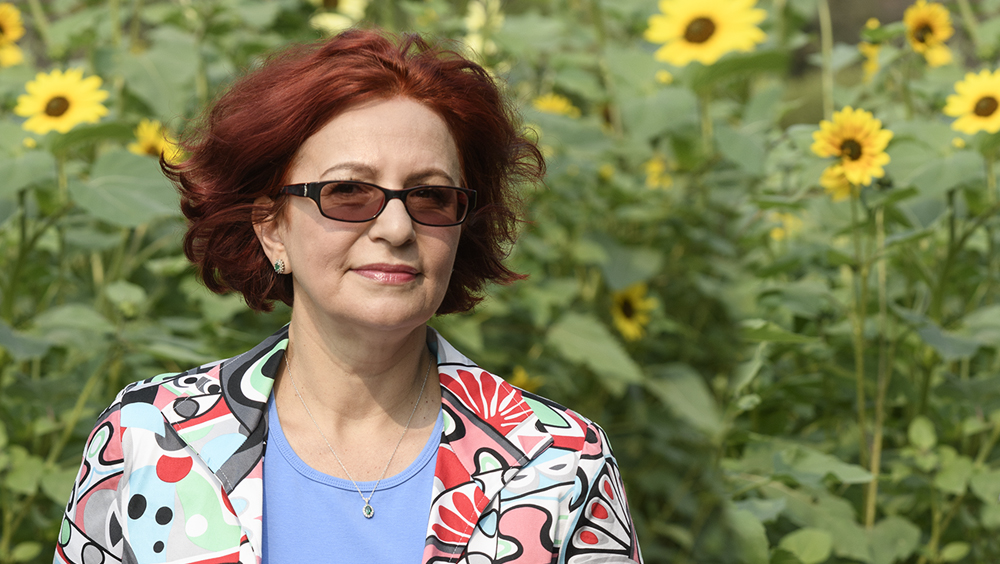 Laura stands in front of a field of sunflowers wearing sunglasses and a patterned shirt. 