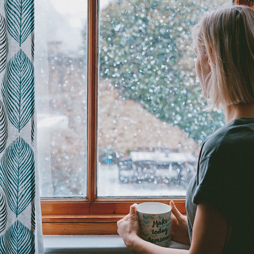 A woman holding a coffee mug standing next to the window and looking outside.