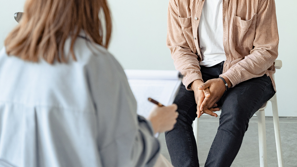 A counselor is seen writing down notes on her clipboard and in the background, her client can be seen seated across her.