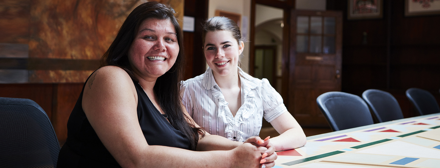 Two smiling women, sitting side by side at a table in a meeting room