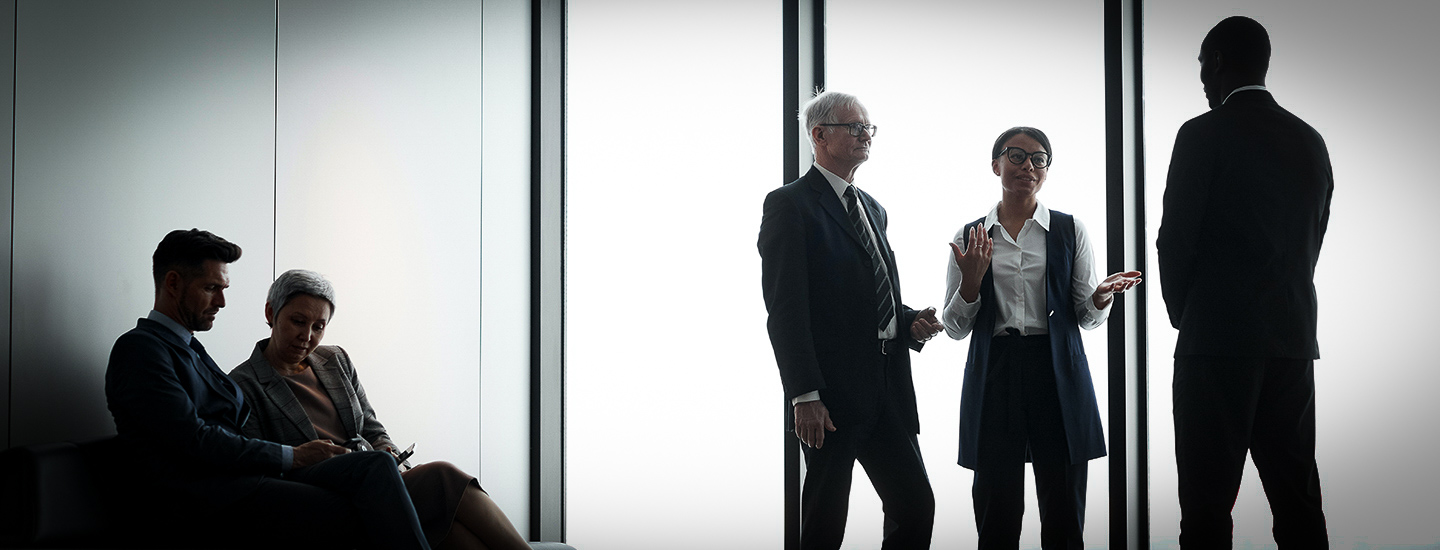 Business people having conversation indoors, three people standing, two people sitting on a chair.