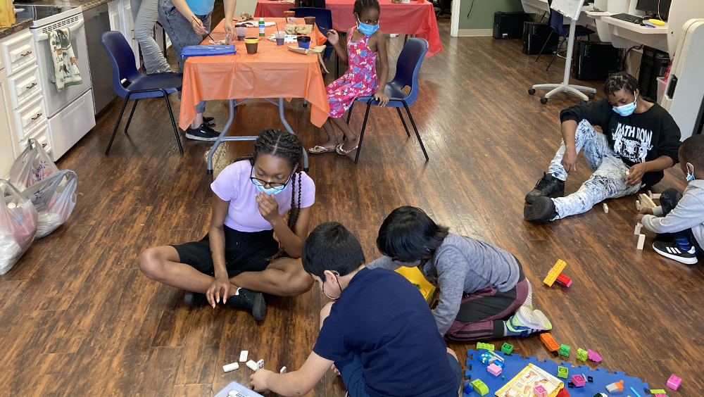 Summer camp instructors helps kids build a structure with blocks on the floor. Little girl with brush painting on table in the back.