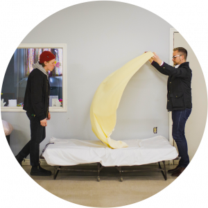 Two men standing in homeless shelter, making bed.