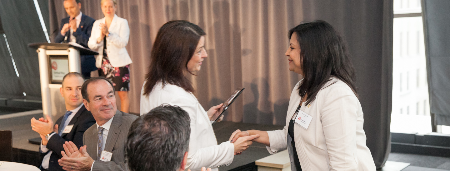 A woman congratulate a female award recipient handshaking and greeting in a event. Group of people applaud in the background.