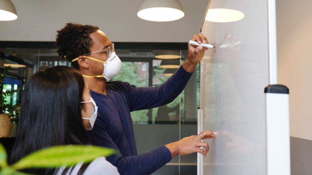 A man wearing a mask writes on a whiteboard as a woman wearing a mask watches