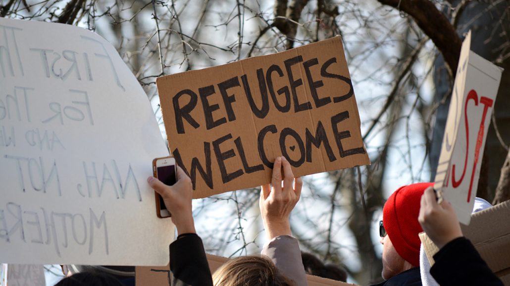 Photograph of people holding up signs. The most prominent says 'Refugees Welcome'