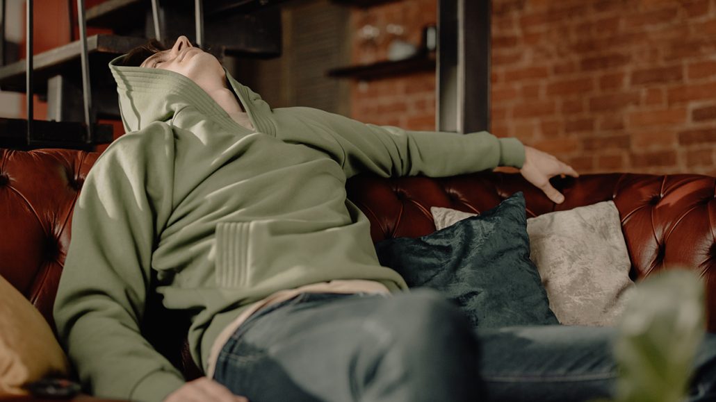 Photograph of young man wearing a hoodie, sitting on a couch and looking up at the ceiling, as if he were defeated and exhausted