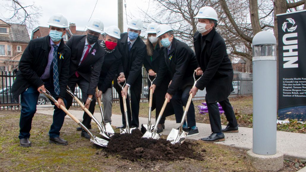 A group of masked speakers wearing suits breaks ground with shovels at the UHN news conference.