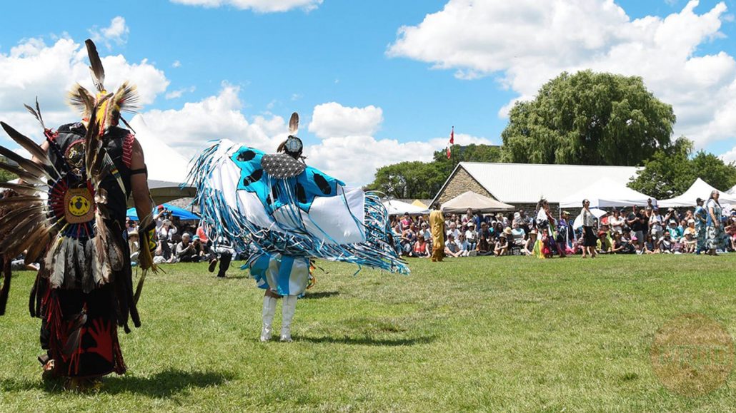 Photo of two Indigenous dancers dressed in regalia performing outside in front of an audience.