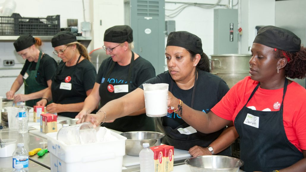Photo of five women dressed in aprons and caps making food in an industrial kitchen