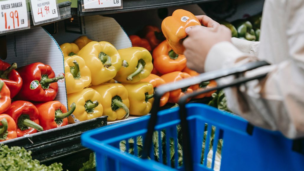 A person at a grocery store holding a basket while picking up a pepper