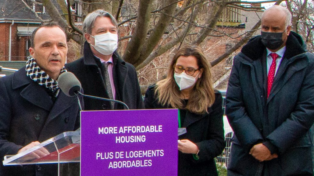 Daniele Zanotti stands at a podium with a sign reading 'More affordable housing' affixed to it. Representatives from different levels of government stand next to him.