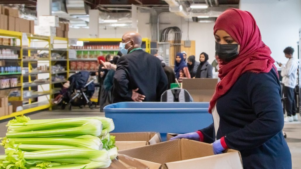 Photo of people wearing masks packing boxes of food at a food bank with a line of people behind them.