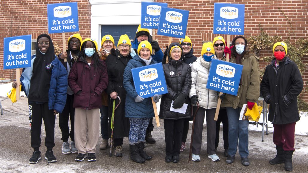 A group of people in yellow beanies with signs that read "Coldest Night: It's cold out here" pose on a snowy sidewalk. 