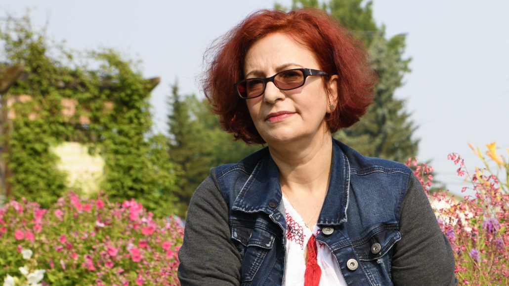 A woman named Laura, with short red hair and wearing glasses and a jean jacket, stands in a garden
