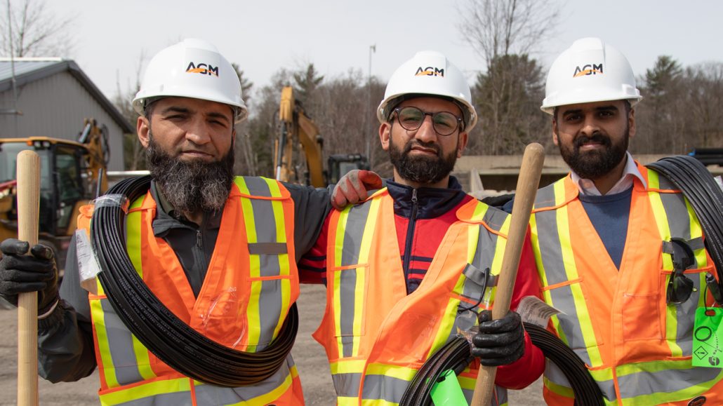 Three construction workers, wearing hardhats and safety vests, look at the camera in front of a construction area.