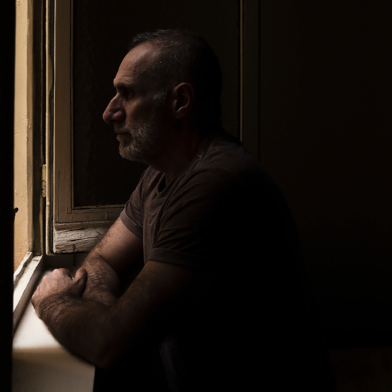 Man alone in a dark room looking out of window.