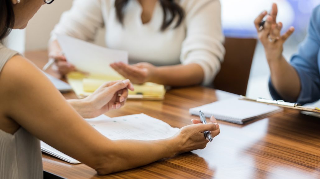 Photo of three women sitting around a conference table working on a project together. 