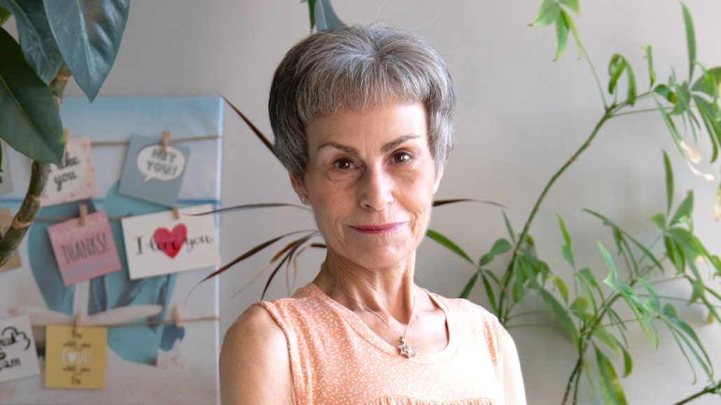 A woman with gray hair wearing a green and white shirt looking directly at the camera and standing in front of a brick wall.