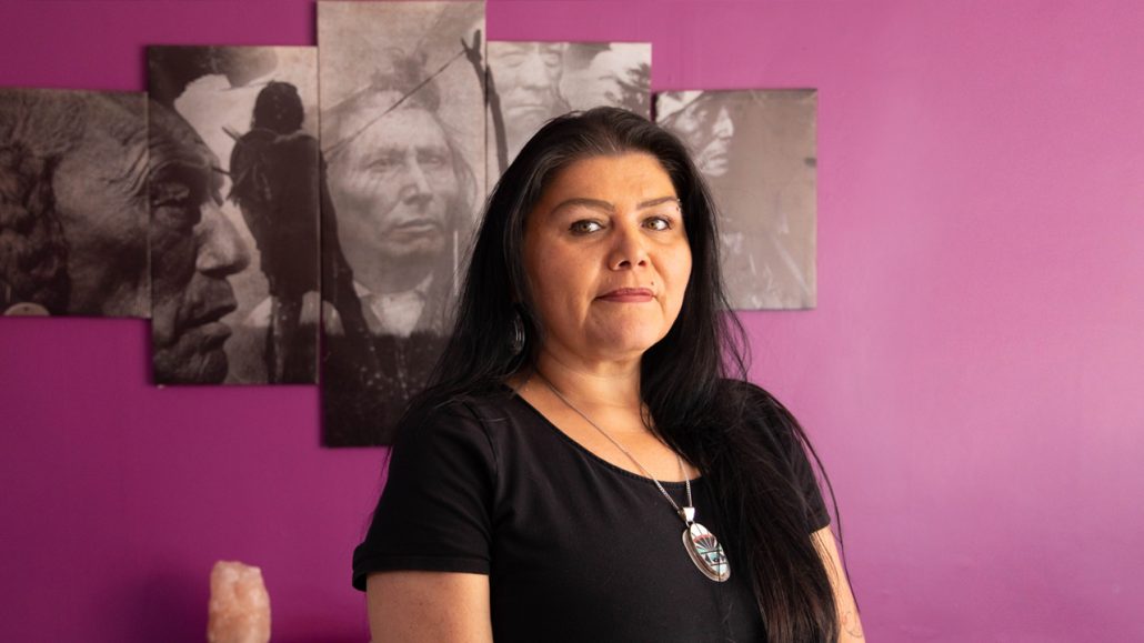 Cynthia Bell, an Indigenous woman wearing a black T-shirt standing in front of a purple wall.
