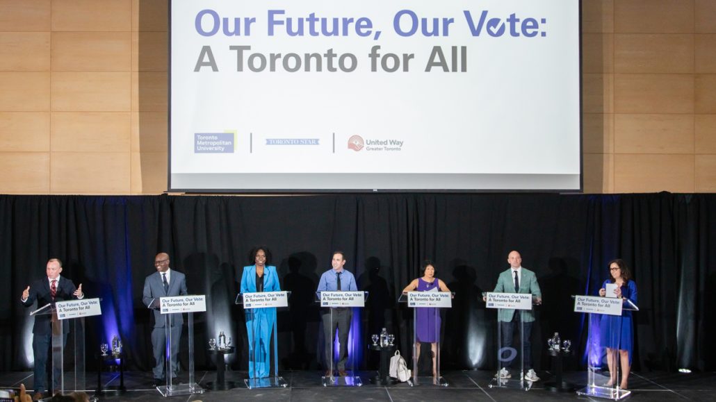 The six leading Toronto mayoral candidates and debate host standing on stage behind podiums in front of a black backdrop at the Toronto For All debate.