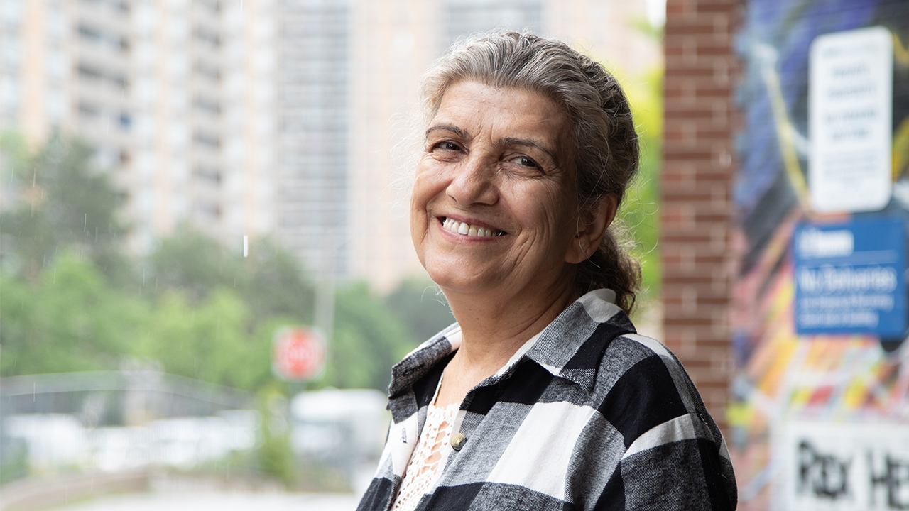 Nafisa smiles at the camera wearing a black-and-white checked top. She's standing in front of a mural and a city street in the summer. 