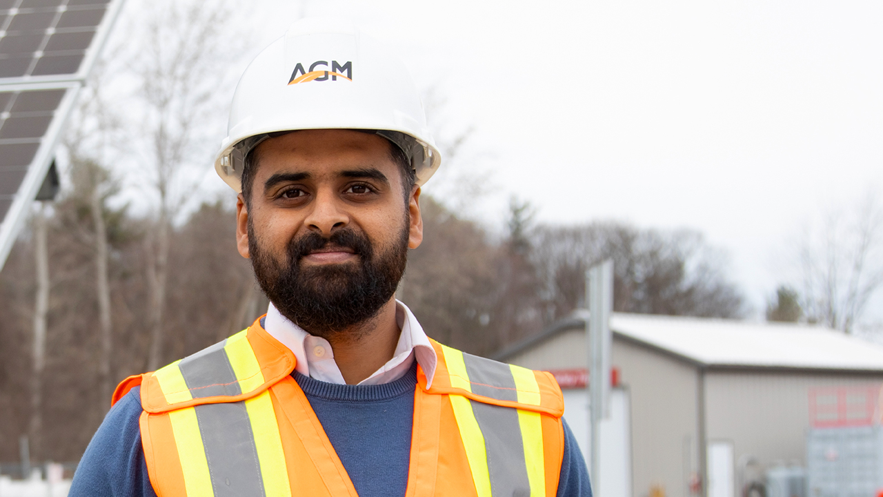 Syed wears a hard hat with "AGM" on it and a orange safety vest and stands in front of a worksite in the winter. 