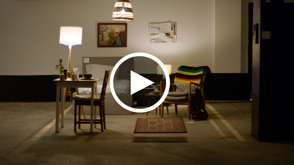 Still of an underground parking space decorated with a bed, kitchen table, chair and bulletin board to look like an apartment.