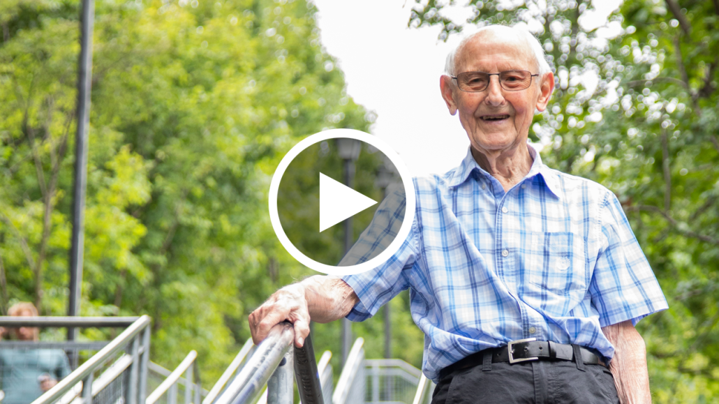 Photo of a senior man in a blue checked shirt smiling at the top of a staircase with green trees in the background.