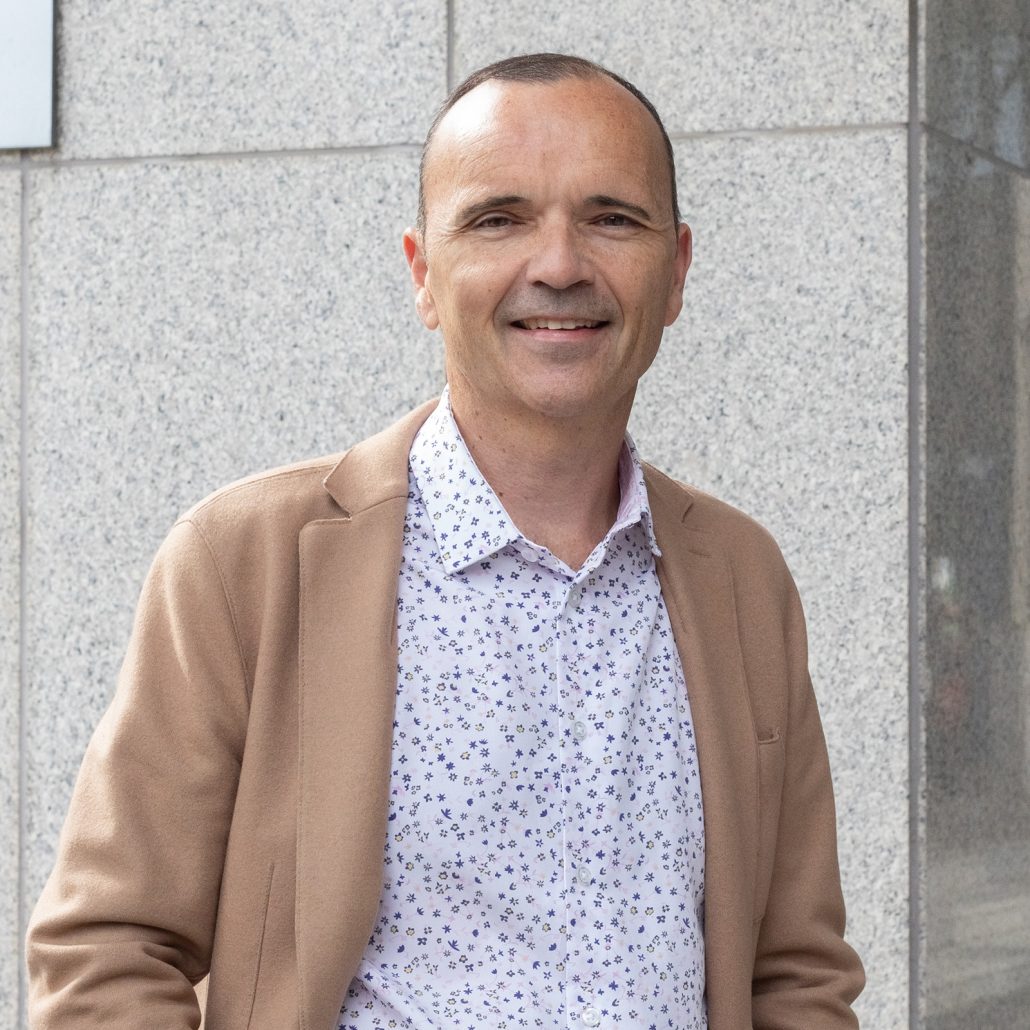 Daniele Zanotti, President and CEO of United Way Greater Toronto, stands in front of a gray wall. He is wearing a white shirt with blue dots and a tan jacket.