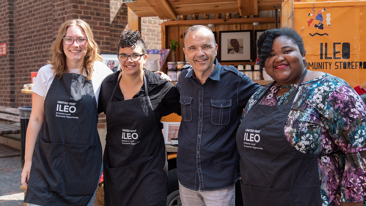 Four people stand in front of a yellow booth with ILEO Community Storefront written in black letters on the side and filled with small jars of products.