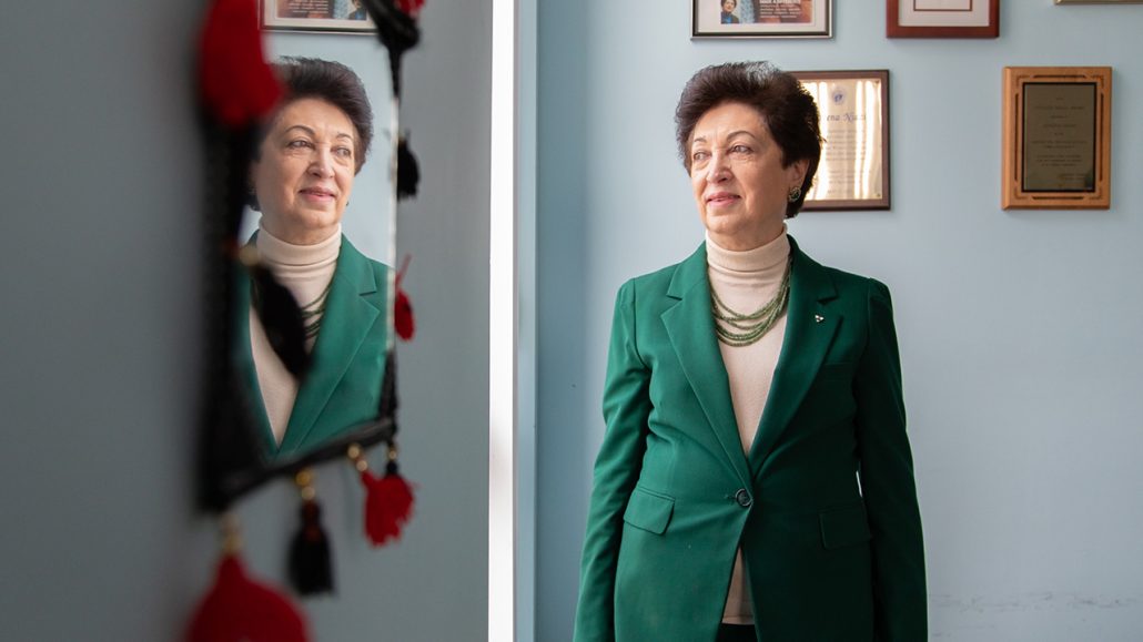 Adeena Niazi, executive director of Afghan Women’s Organization Refugee and Immigrant Services, stands with her arms crossed in front of an evergreen tree. She has short brown hair and is wearing a white turtleneck and green blazer.