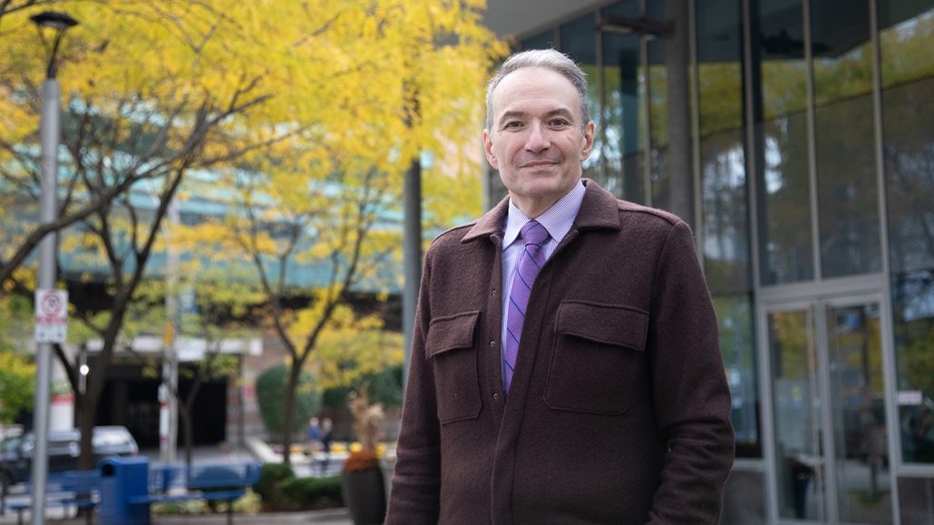 Kevin Smith, University Health Network’s CEO, stands outside looking at the camera. He has grey hair and is wearing a burgundy jacket, button down shirt and purple and blue striped tie.
