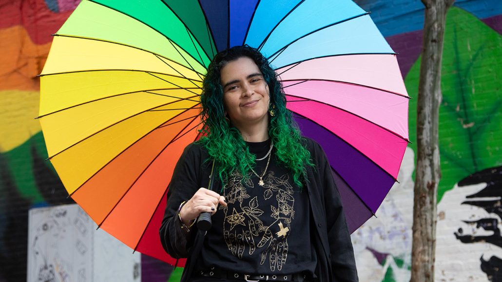 Eli, who got support at The 519, a United Way-funded organization that supports the LGBTQ+ community, poses in front of an urban mural holding a rainbow umbrella.
