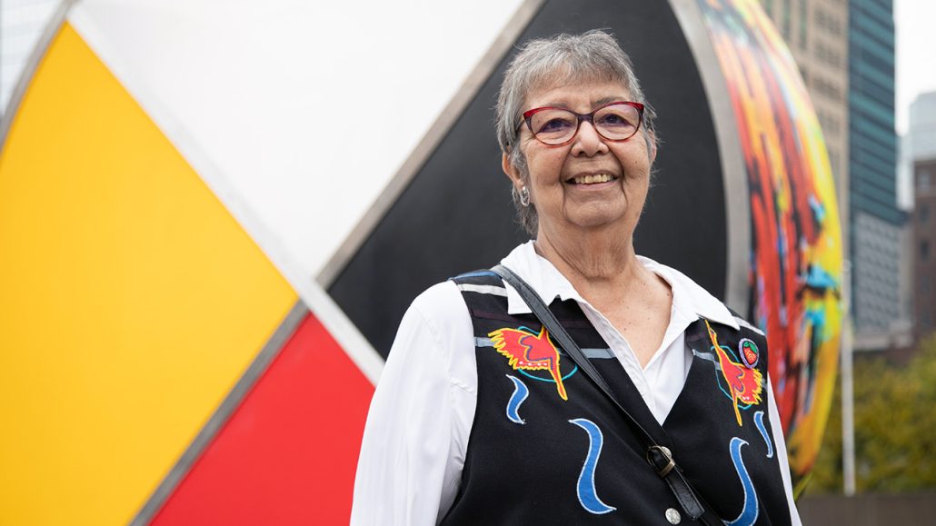 Frances Sanderson, executive director of Nishnawbe Homes, stands in front of a medicine wheel sign smiling at the camera. She has short grey hair and is wearing red framed eyeglasses. 