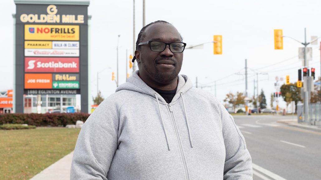 Bryan Peart wears a grey sweatshirt and glasses and stands at an intersection in front of sign that reads "Golden Mile"