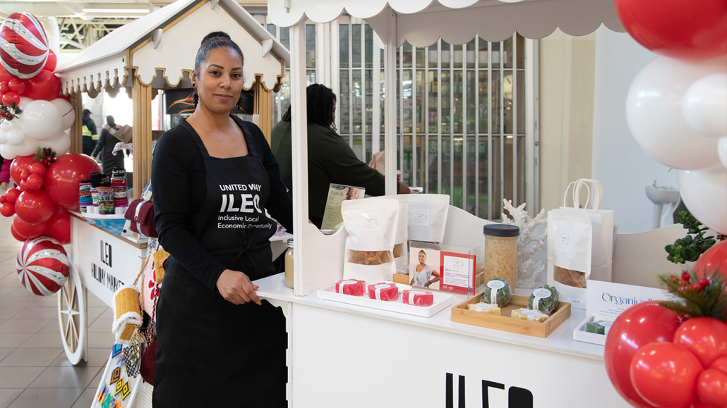 Vendor Raven Joyce stands with her wares displayed on a festive white wagon-shaped table, decorated with red and white balloons, at the holiday market.