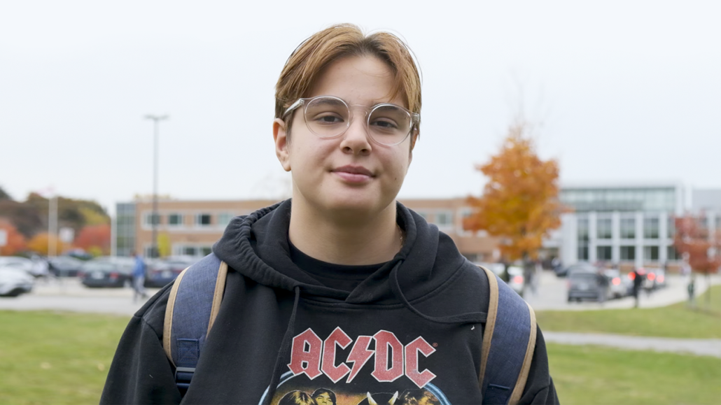 Serpil is wearing clear framed glasses, a black AC/DC hoodie and a backpack. She stands looking at the camera with a high school blurred in the background.