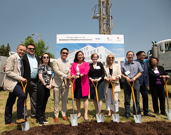 Representatives from United Way Greater Toronto, Scarborough Health Network, YMCA of Greater Toronto and government pose at a groundbreaking event. They are holding shovels by a mound of dirt, in front of a sign that says “Future Home of the Bridletowne Neighbourhood Centre”.