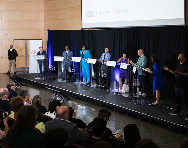 The six leading Toronto mayoral candidates and debate host standing on stage behind podiums in front of a black backdrop at the Toronto For All debate.