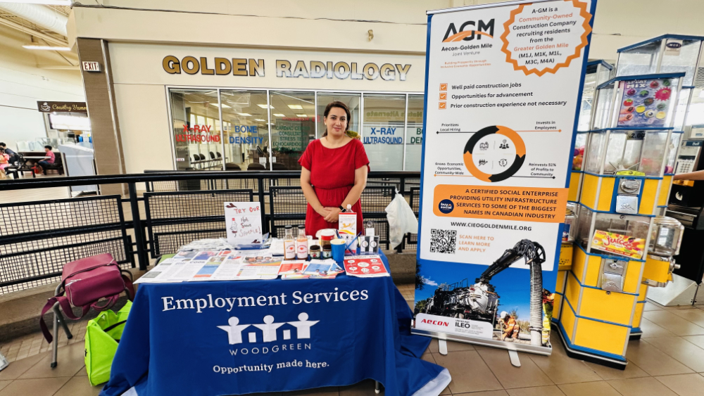 A woman in a red dress representating community organization Woodgreen stands behind a table that features information about their employment services work.