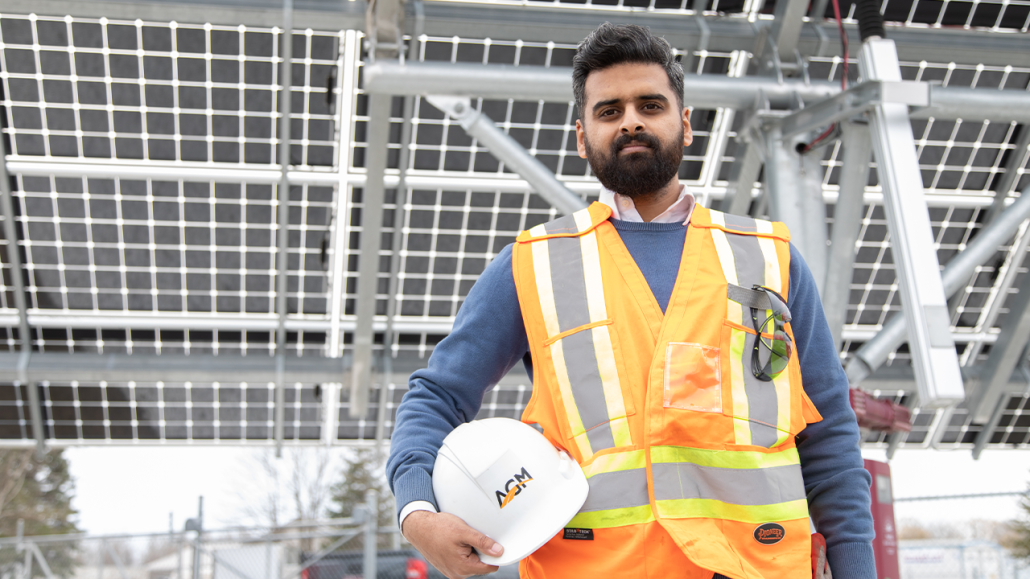 Syed wears a safety vest and holds a hardhat while standing in front of a jobsite.