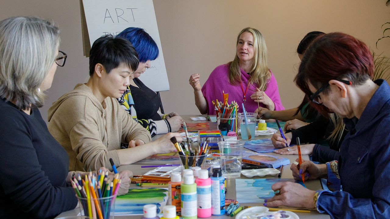 A group of people sit at a table filled with paints, pencil crayons and other art supplies.