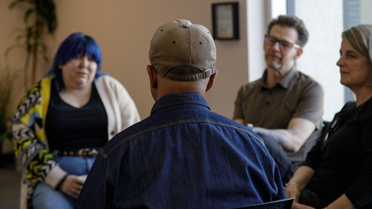 A man is photographed from behind. Blurred in the background are people sitting in a circle listening to the man speak.